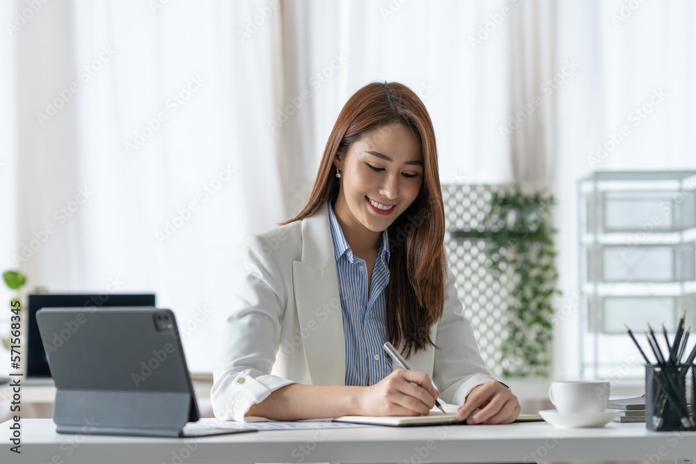 Confident Asian businesswoman sitting and taking notes in financial book Income tax with laptop computer or tablet in a happy office. Confident Asian businesswoman sitting and taking notes in financial book Income tax with laptop computer or tablet in a happy office.