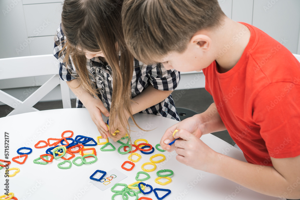 Little school kids play toy constructor standing above kitchen table and looking down, from above view. Boy and girl collect plastic colored geometric figures. Entertaining interesting board game. Little school kids play toy constructor standing above kitchen table and looking down, from above view. Boy and girl collect plastic colored geometric figures. Entertaining interesting board game.