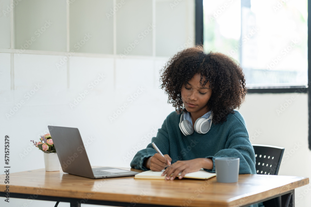 Cute african american female student wearing headphones with afro dreadlocks, studying remotely from home, using a laptop, and taking notes on a notepad online. Cute african american female student wearing headphones with afro dreadlocks, studying remotely from home, using a laptop, and taking notes on a notepad online.