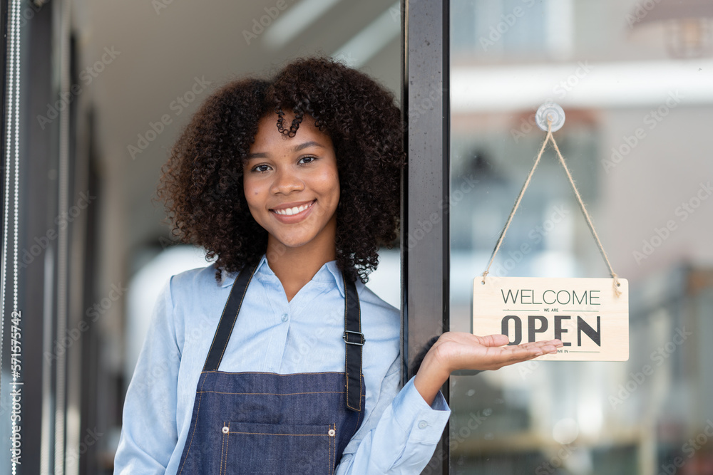 Φωτογραφία African young woman starting a small business working in a cafe A coffee shop worker stands in front of the store opening an open sign and greets customers with happy smiles