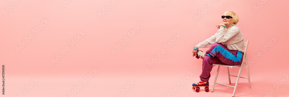 Beautiful old woman, grandmother in stylish sportive trousers and sunglasses posing on vintage rollers over pink studio background. Age, fashion, lifestyle, emotions concept. Banner. Copy space for ad - obrazy, fototapety, plakaty 