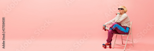 Beautiful old woman, grandmother in stylish sportive trousers and sunglasses posing on vintage rollers over pink studio background. Age, fashion, lifestyle, emotions concept. Banner. Copy space for ad