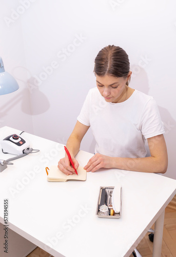 Nail technician makes notes in notebook with pen, sitting at workstation, table in nail beauty treatment salon. Professional tools are on white table. Manicurist's routine. Vertical, closeup photo