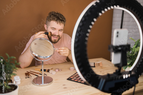 Portrait of happy man beauty blogger sitting in room at table and speaking recommending foundation or decorative cosmetic looking at camera. Blogging and vlog concept