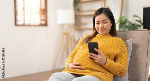 Obraz na plátně Happy Asian pregnant woman using her phone while relaxing on sofa in her living