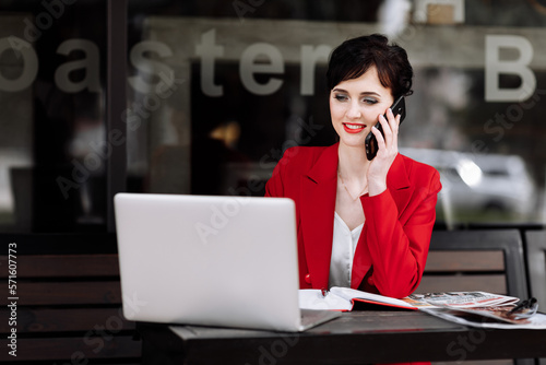 Smiling businesswoman talking on the smartphone, working on a laptop and doing notes at cafe on terrace in urban city. Office worker communication with clients remote. Coffee break, business concept
