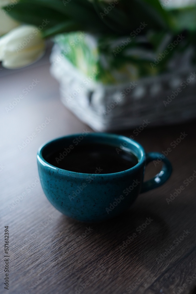 A close-up of a turquoise cup of black coffee against a basket of white tulips. A beautiful blue cup of hot coffee sits on a wooden table near the window A close-up of a turquoise cup of black coffee against a basket of white tulips. A beautiful blue cup of hot coffee sits on a wooden table near the window