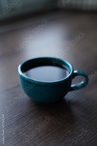 A close-up of a turquoise cup with black coffee against a wooden table. A beautiful blue cup of hot coffee sits on a wooden table near the window