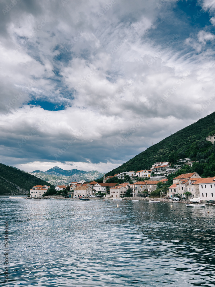 Coast of Perast with ancient stone houses near the green mountains against the background of a cloudy sky