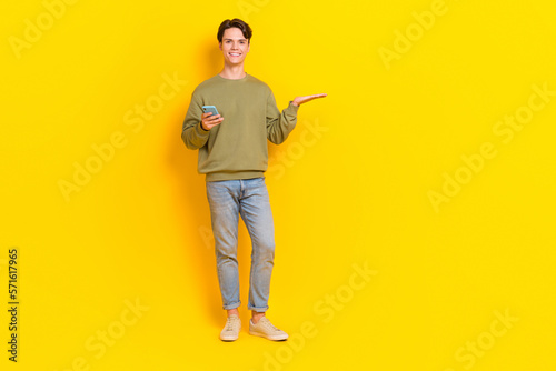 Full body photo of positive young man hold smart phone arm palm demonstrate empty space isolated on yellow color background