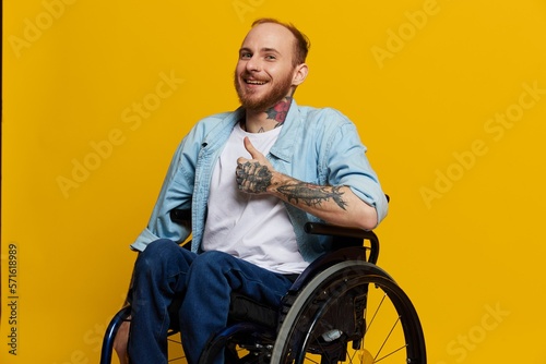 A man in a wheelchair smile and happiness, thumb up, with tattoos on his hands sits on a yellow studio background, the concept of health a person with disabilities