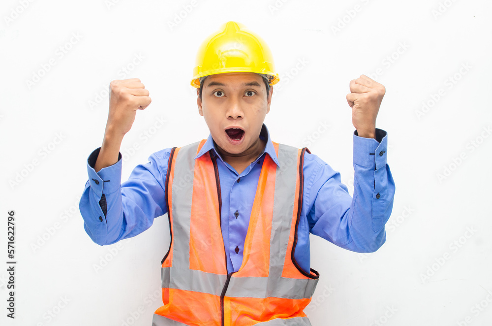 overjoyed asian male construction worker in blue shirt and orange vest with yellow safety helmet doing yes gesture by raising his fist.
