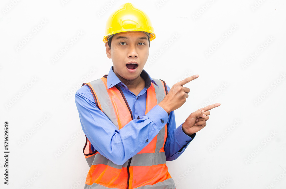 asian construction worker wearing yellow safety helmet and vest smiling and pointing finger with happy and shocked expression. billboard model advertisment concept. 