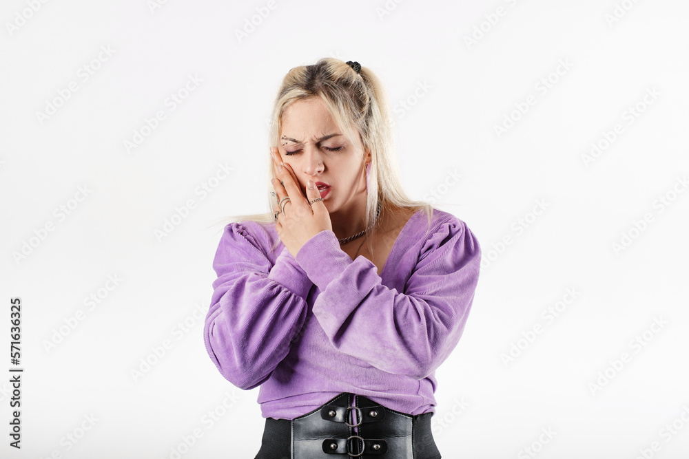Young blonde woman wearing lilac ribbed blouse isolated over white background with mouth open, touching cheek with both hands, is suffering from a toothache. Looks very sad. Young blonde woman wearing lilac ribbed blouse isolated over white background with mouth open, touching cheek with both hands, is suffering from a toothache. Looks very sad.