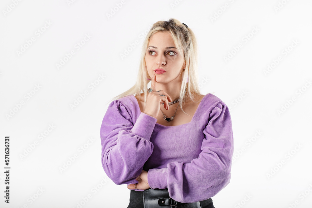 Young beautiful woman wearing lilac ribbed blouse isolated over white background undecided, purses lips and touches with index finger. Looks at the empty copy space and tries to decide. Young beautiful woman wearing lilac ribbed blouse isolated over white background undecided, purses lips and touches with index finger. Looks at the empty copy space and tries to decide.
