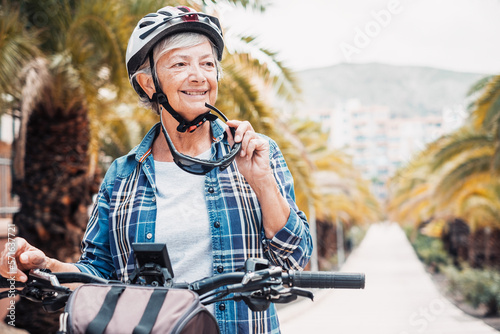 Portrait of attractive senior woman with helmet riding her electric bicycle in urban park. Elderly smiling lady enjoying healthy lifestyle, vacation, retirement