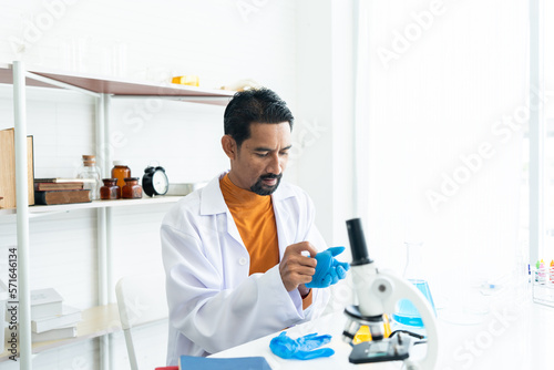 A male teacher in white lab coat wearing rubber gloves  preparing many laboratory tools shelves with microscope  textbooks  yellow and blue chemical flasks on table in university science classroom.