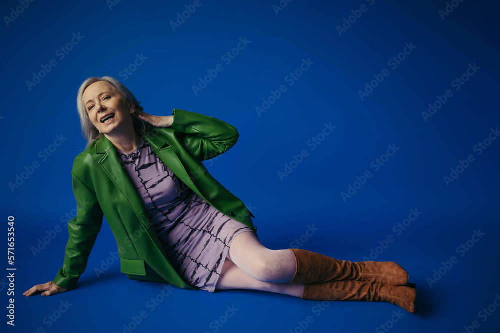 full length of cheerful senior lady in green leather jacket and purple dress sitting on blue background