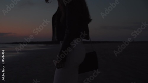 Young attractive girl wearing white jeans walks on the sand of a beach at sunset in Chipiona, Cadiz, Spain. Shot of her hips and bottom with the sea and the sky on the background. She has a purse too. photo