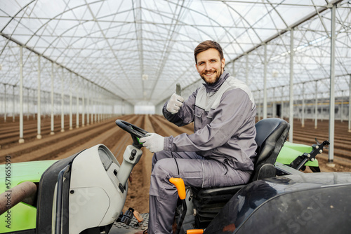 An agronomy worker is driving tractor in hothouse and giving thumbs up while smiling at the camera.