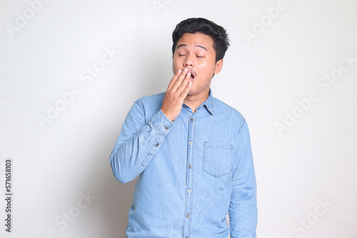 Portrait of sleepy Asian man in blue shirt yawning with closed eyes due to overworking while covering mouth with hand. Isolated image on white background