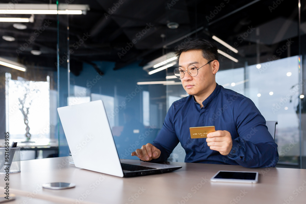A young male Asian banker works in the office at a laptop, holds a credit problem in his hands. Advises, talks, offers an account via video call to clients. A young male Asian banker works in the office at a laptop, holds a credit problem in his hands. Advises, talks, offers an account via video call to clients.