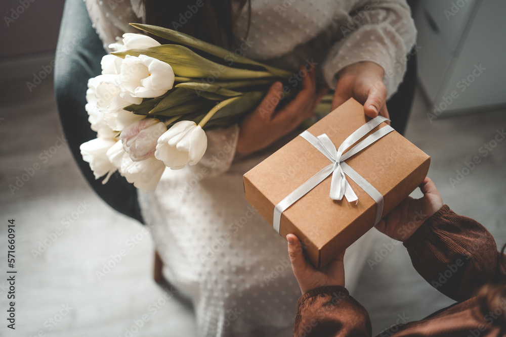 Obraz premium The child's hands hold a beautiful gift box with a ribbon and white tulips. Top view, close-up. Happy mother's day