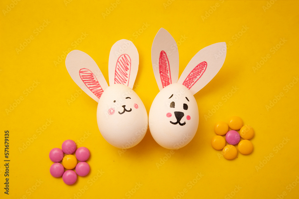 Food photo. White chicken egg with cute bunny faces and bunny ears. Postcard for religious holiday. Small round multicolored bright candies in form of flowers on yellow background. happy easter. Food photo. White chicken egg with cute bunny faces and bunny ears. Postcard for religious holiday. Small round multicolored bright candies in form of flowers on yellow background. happy easter.