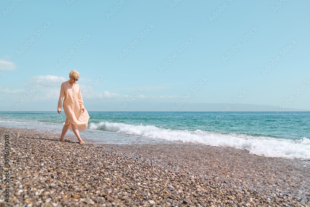 Pretty blond woman in sunglasses and transparent dress walking in the beach, smiling and enjoying sunny windy day at the sea. Travel concept. Pastel muted color. Pretty blond woman in sunglasses and transparent dress walking in the beach, smiling and enjoying sunny windy day at the sea. Travel concept. Pastel muted color.