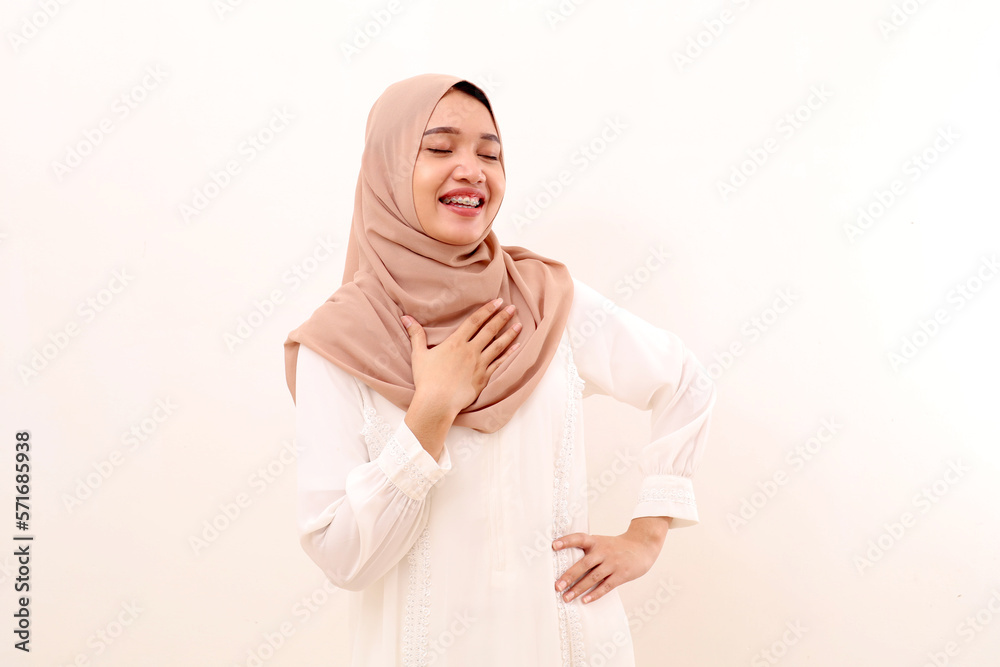 Portrait of young asian muslim woman showing relieved gesture while holding chest and closing eyes