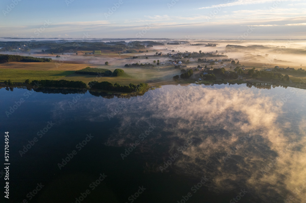 Foggy morning on a beautiful lake surrounded by forests and fields. A small village located near a lake in fog and morning sun.