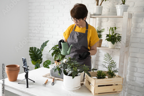 Middle aged woman blogger in front of smartphone camera on tripod records instructional tutorial video for her blog shoots process of replanting flowers and green plants full of soil enjoys botanic