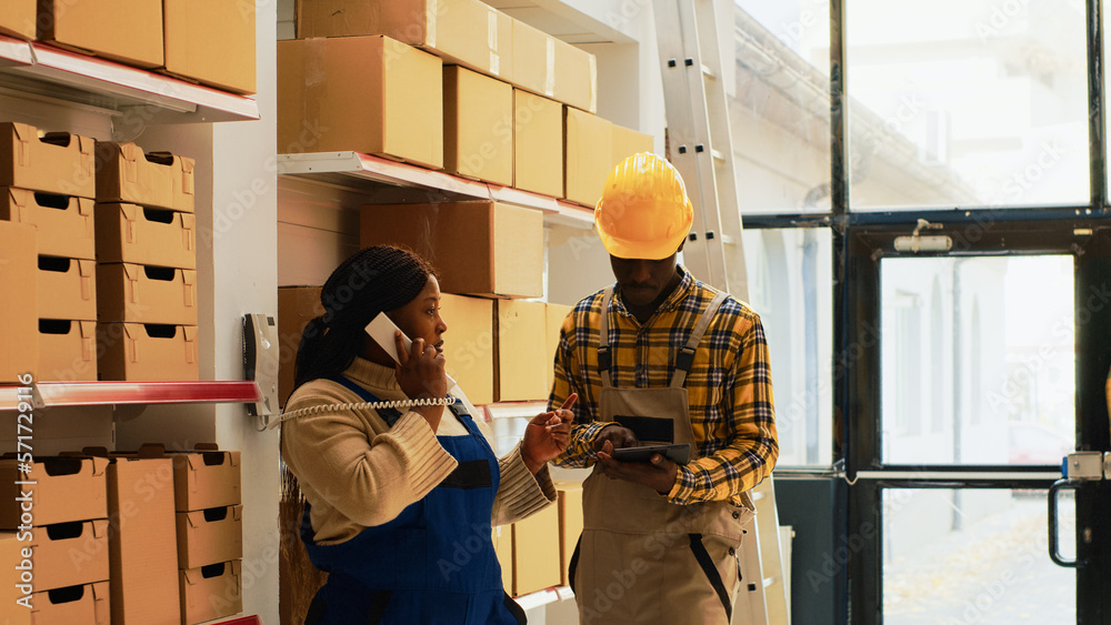 Depot manager answering phone call in warehouse space, preparing new supplies order with male employee. Young man writing list of goods on clipboard files, supervisor using landline phone. Depot manager answering phone call in warehouse space, preparing new supplies order with male employee. Young man writing list of goods on clipboard files, supervisor using landline phone.