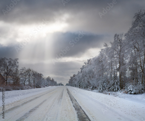 Winter landscape with ice-covered road, trees at side of the road, and sunrays from cloudy sky.