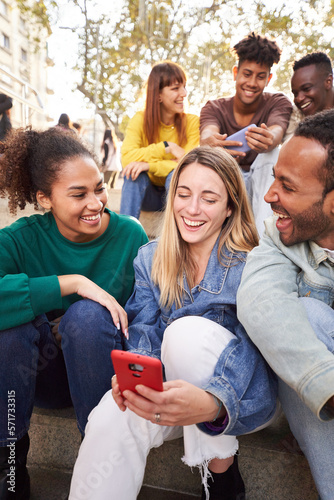 Vertical photo of group of young people looking at and using phone sitting on stairs. Smiling multicultural friends looking for information outside the university campus. Students in their free time.