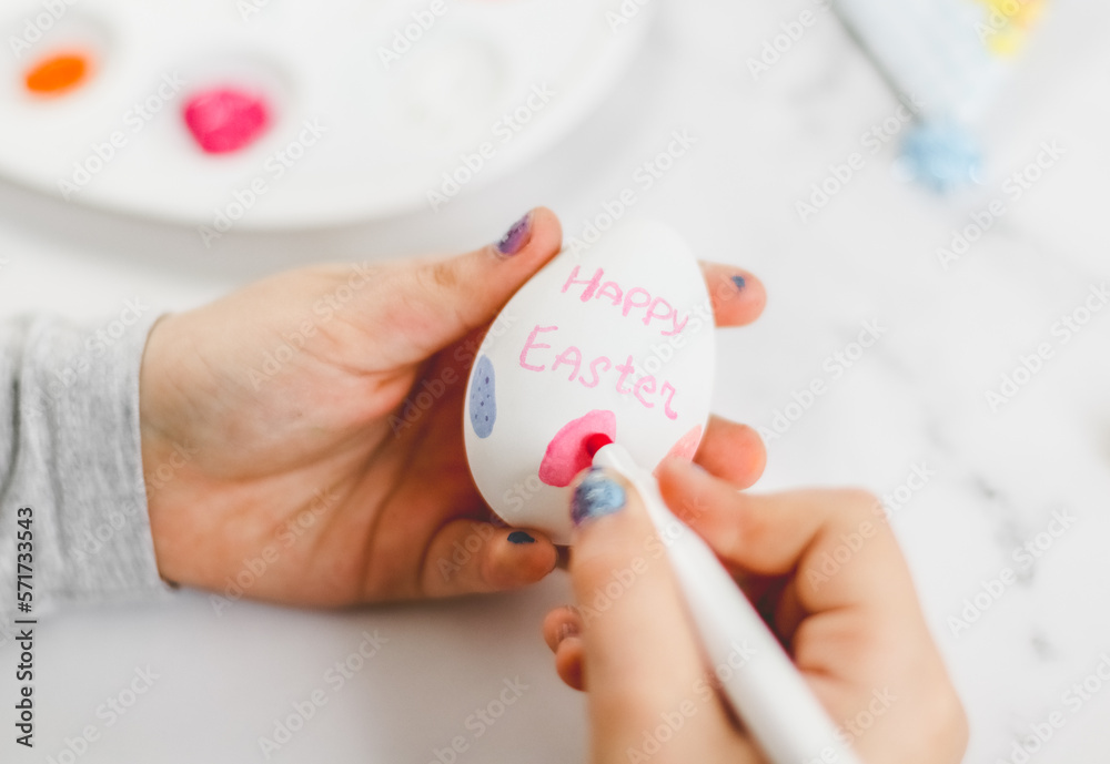 The hands of a caucasian little girl hold an egg and draw on it with a pink marker Happy Easter The hands of a caucasian little girl hold an egg and draw on it with a pink marker Happy Easter