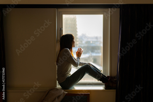 Loneliness Asian woman sitting by the window in bedroom and looking cityscape building covered in snow at winter sunset. Attractive girl resting in hotel room during travel alone on holiday vacation.