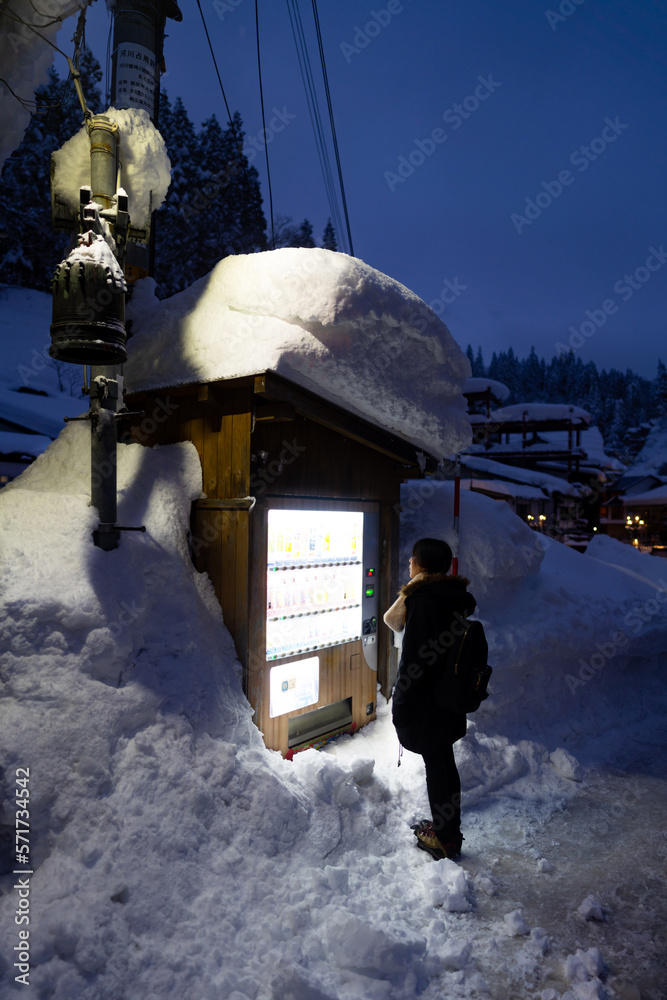 Fototapeta premium Asian girl in winter coat choosing and buying hot drinks in beverage vending machine covered in snow during travel small town at night. Young woman travel local village in Japan on winter vacation.