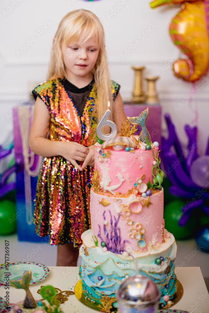 Indoor shot of a rather joyful little girl with blond hair making a wish before blowing out the candle. Celebrates her 6th birthday in a trendy dress, with a huge nautical-themed cake. Happy childhood