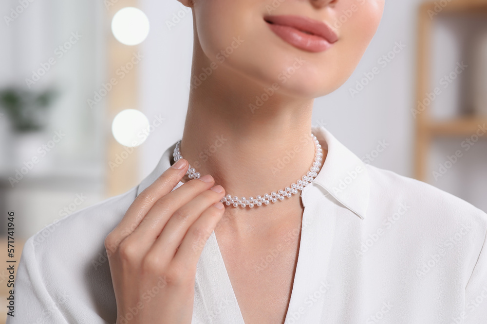 Young woman trying on elegant pearl necklace indoors, closeup