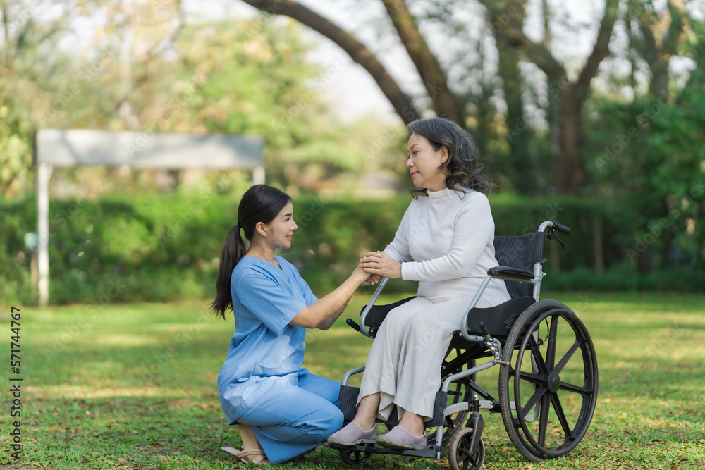 Young asian care helper with asia elderly woman on wheelchair relax together park outdoors to help and encourage and rest your mind with green nature. hold hands to encourage Young asian care helper with asia elderly woman on wheelchair relax together park outdoors to help and encourage and rest your mind with green nature. hold hands to encourage