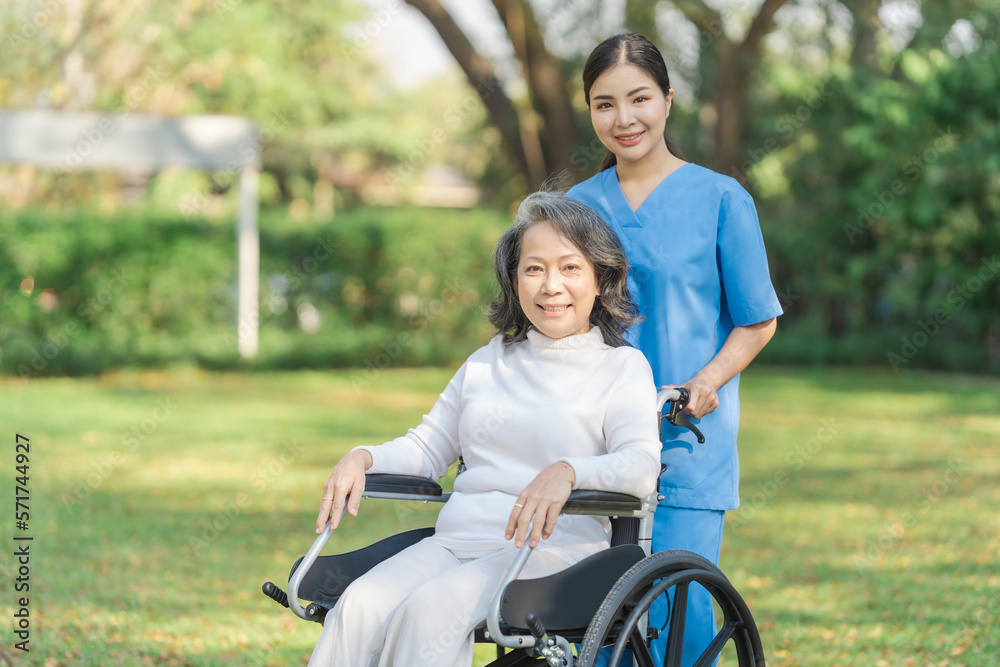 Young asian care helper with asia elderly woman on wheelchair relax together park outdoors to help and encourage and rest your mind with green nature. Looking camera. Young asian care helper with asia elderly woman on wheelchair relax together park outdoors to help and encourage and rest your mind with green nature. Looking camera.