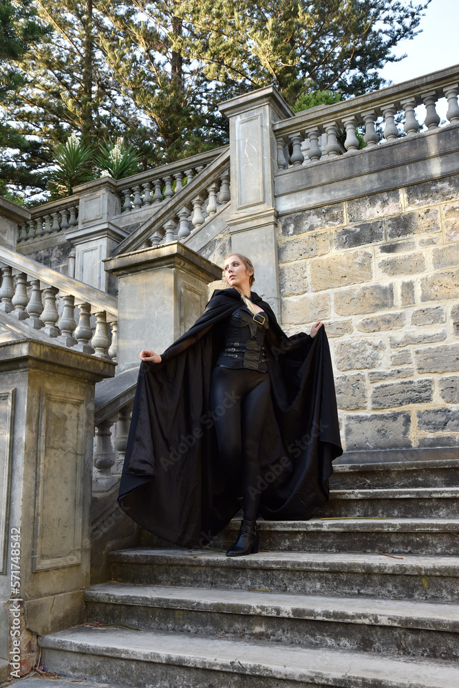 Portrait of beautiful female model with blonde plait, wearing black leather catsuit and flowing hooded cloak, fantasy assassin warrior.  Posing in castle background with stone staircase.