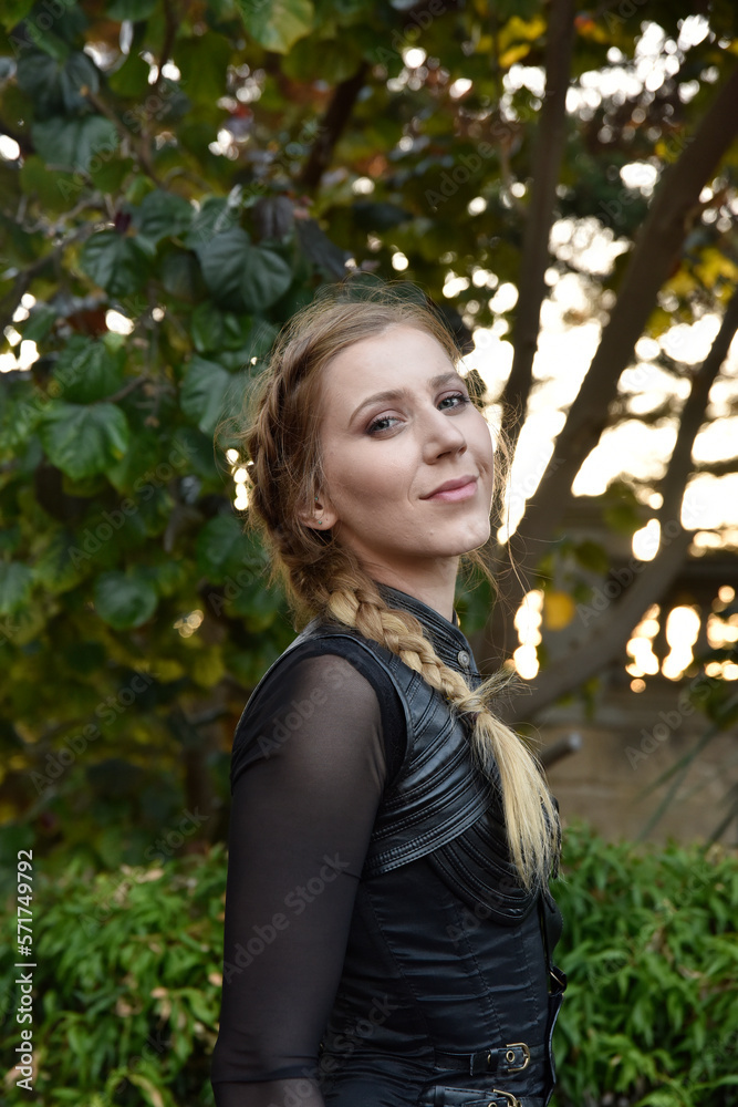Close up portrait of beautiful female model with blonde plait, wearing black leather catsuit, fantasy assassin warrior. Posing in forest tree background Close up portrait of beautiful female model with blonde plait, wearing black leather catsuit, fantasy assassin warrior. Posing in forest tree background
