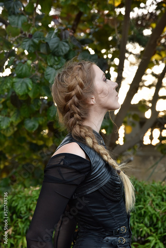 Close up portrait of beautiful female model with blonde plait, wearing black leather catsuit, fantasy assassin warrior.  Posing in forest  tree background