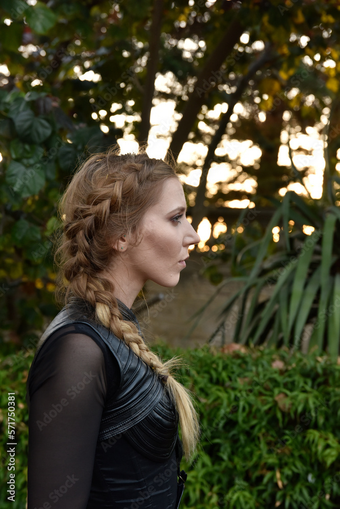 Close up portrait of beautiful female model with blonde plait, wearing black leather catsuit, fantasy assassin warrior. Posing in forest tree background Close up portrait of beautiful female model with blonde plait, wearing black leather catsuit, fantasy assassin warrior. Posing in forest tree background