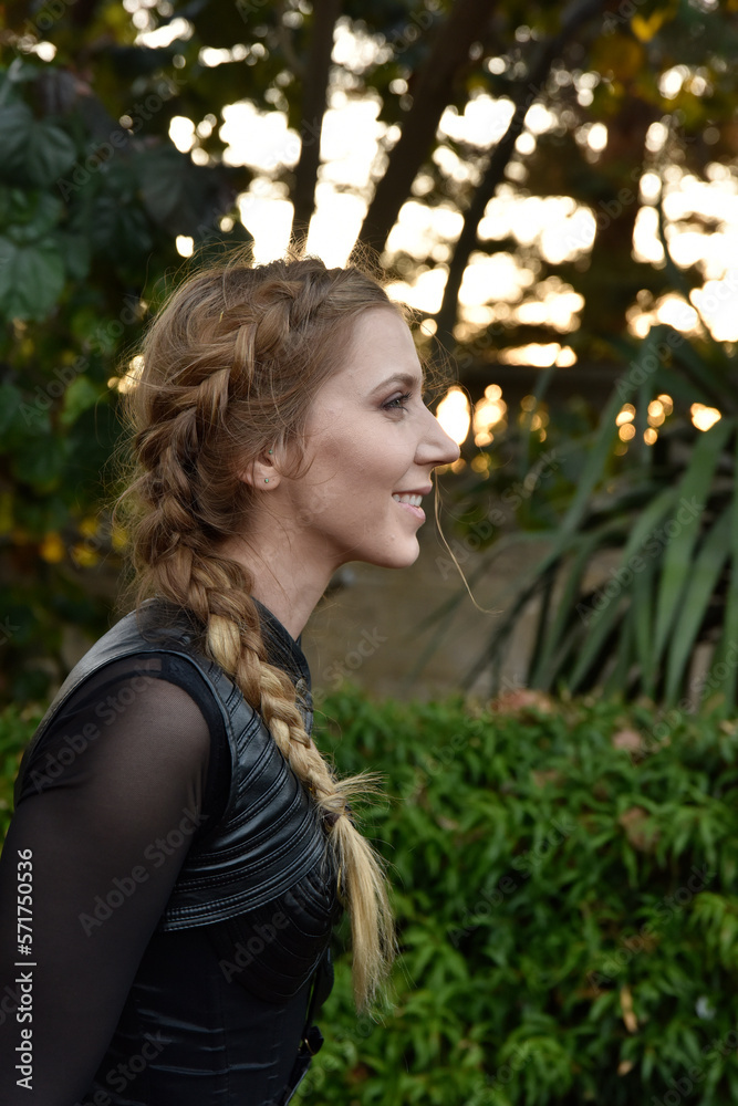 Close up portrait of beautiful female model with blonde plait, wearing black leather catsuit, fantasy assassin warrior. Posing in forest tree background Close up portrait of beautiful female model with blonde plait, wearing black leather catsuit, fantasy assassin warrior. Posing in forest tree background