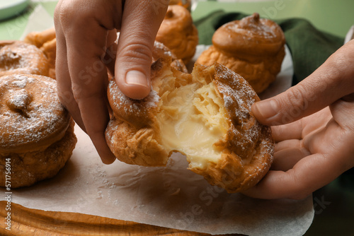 Woman breaking delicious profiterole filled with cream above wooden board, closeup