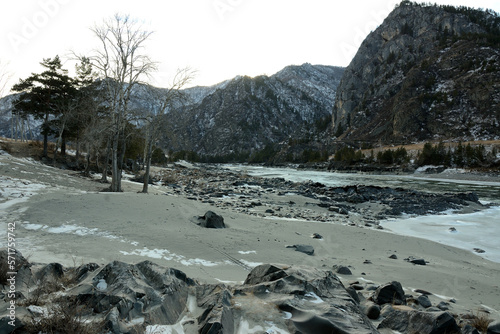 Sandy and rocky shore with lonely trees on the banks of a beautiful frozen river surrounded by snow-capped mountains on a winter evening.