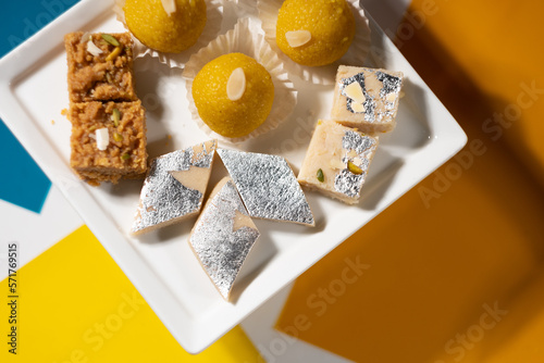 An assortment of traditional Indian desserts (round 'laddu', square beige 'mawa burfi', square brown 'mohanthal', and diamond 'kaju') served on a square white ceramic plate. photo